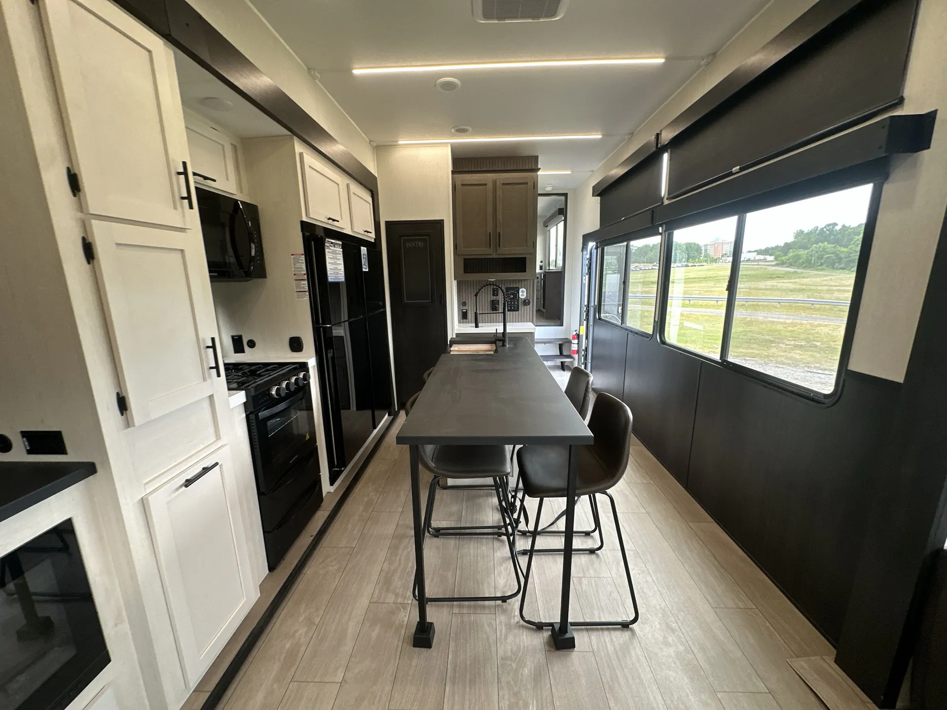 2026 Forest River Arctic Wolf 38LEAH.G — Long dining table with chairs stands centrally between a full kitchen appliance wall and large windows