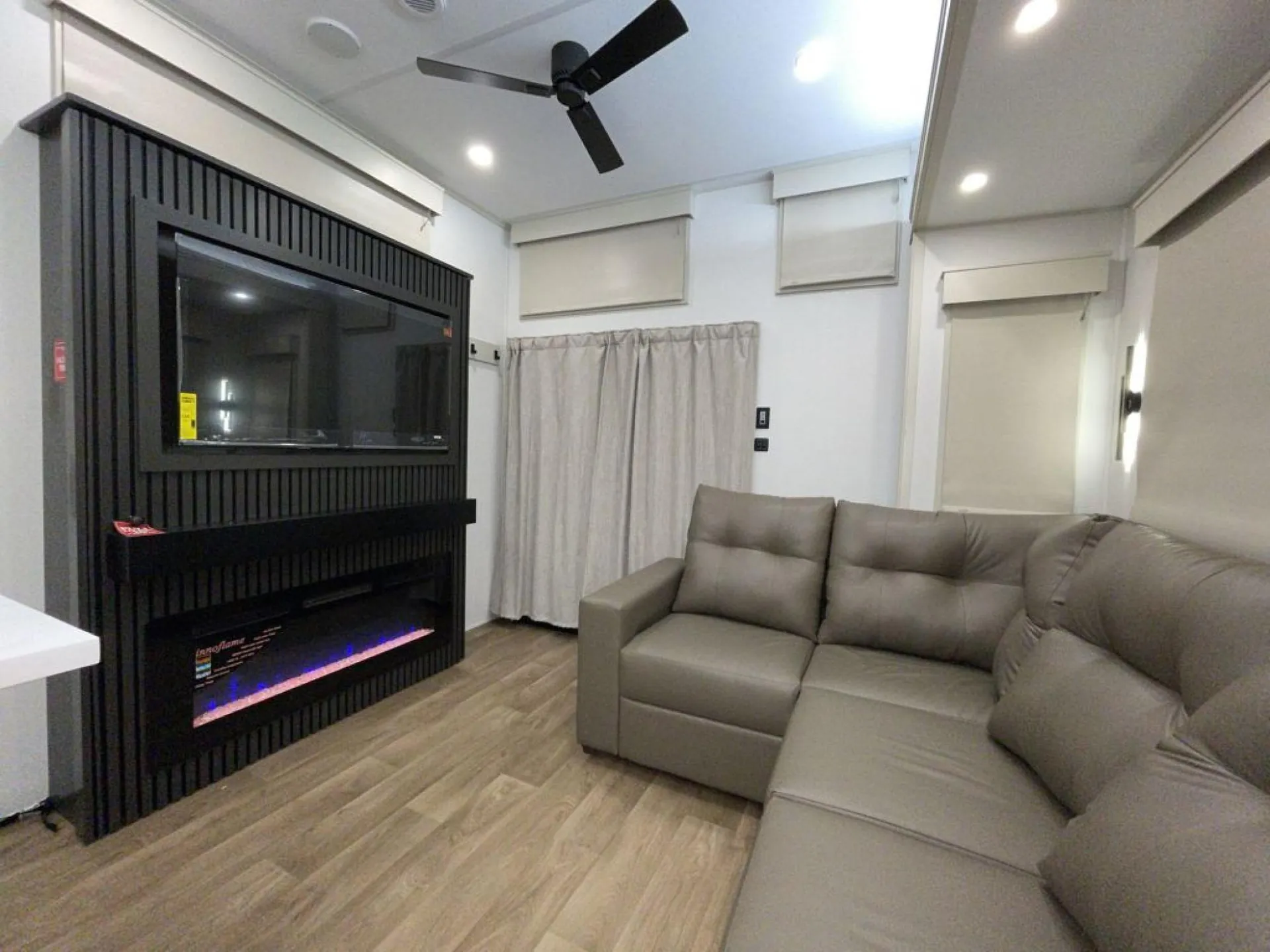 2026 Grand Design Foundation 42GD — Destination trailer living room with sectional sofa, wall-mounted television, electric fireplace, windows, and curtains