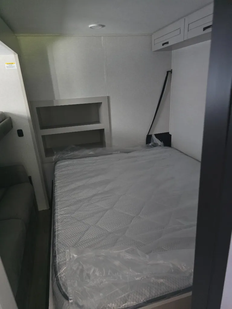2026 Jayco North Point 377RLBH — Main bedroom with a plastic-wrapped bed, recessed bedside shelving, and overhead cabinets
