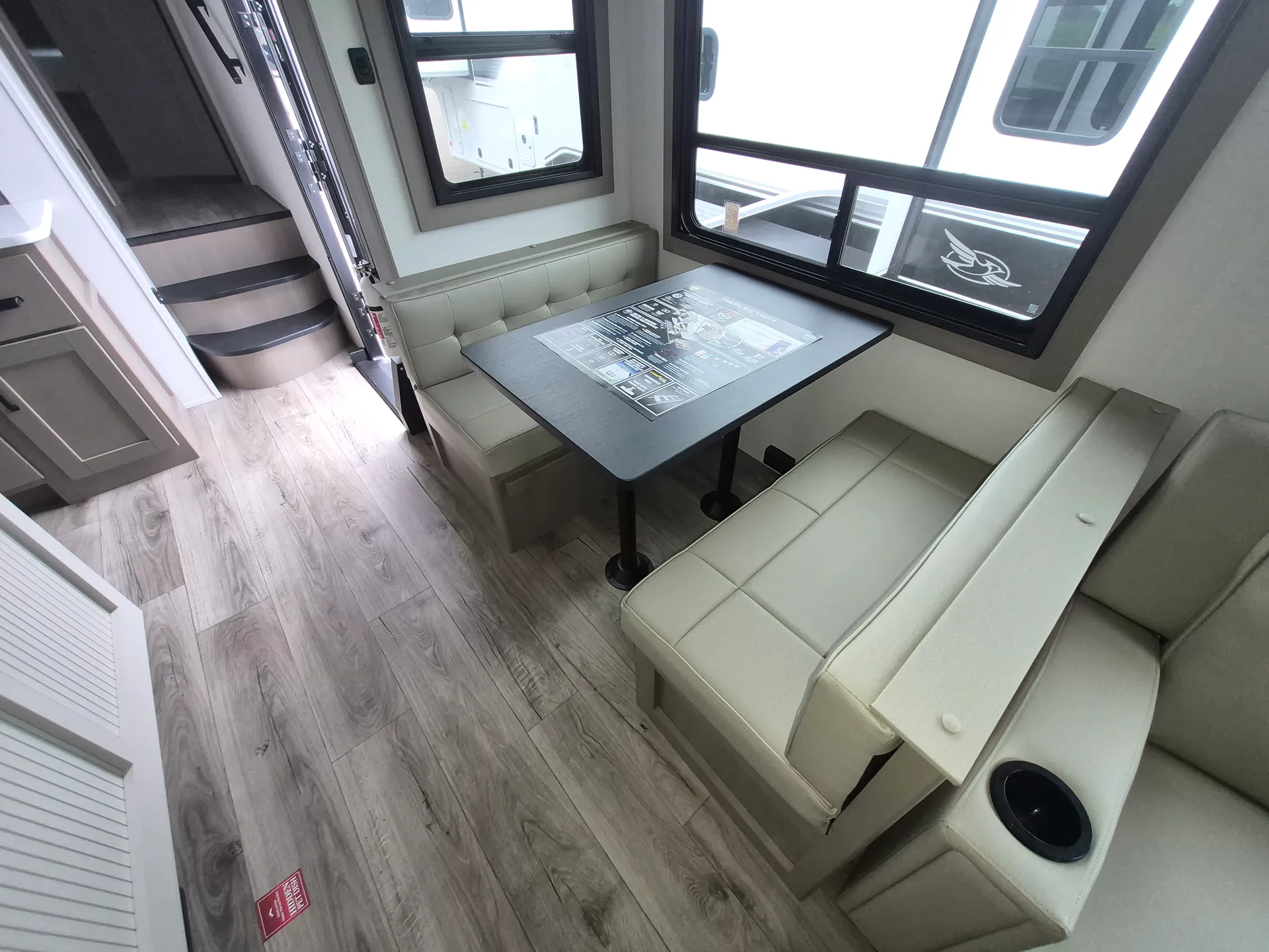 2027 Grand Design Reflection 362TBS — Dinette booth and table beside a sofa, with large windows and a kitchen passage visible behind