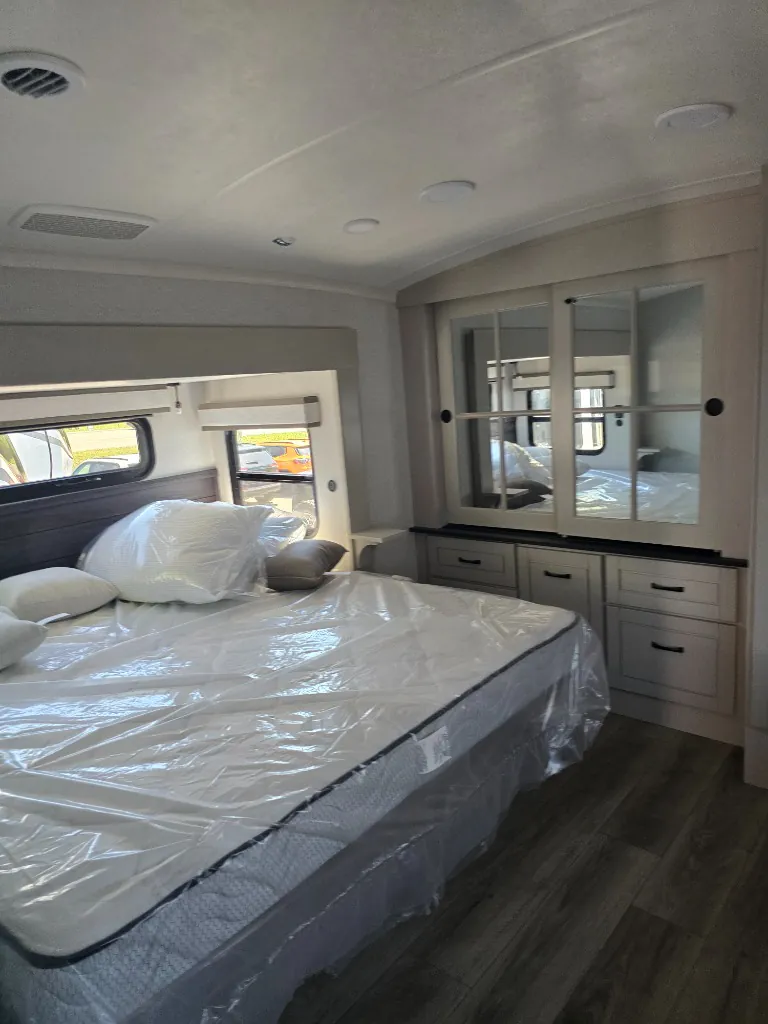 2026 Jayco North Point 377RLBH — Main bedroom with covered bed, pillows, windows, mirrored dresser, and overhead ceiling fixtures