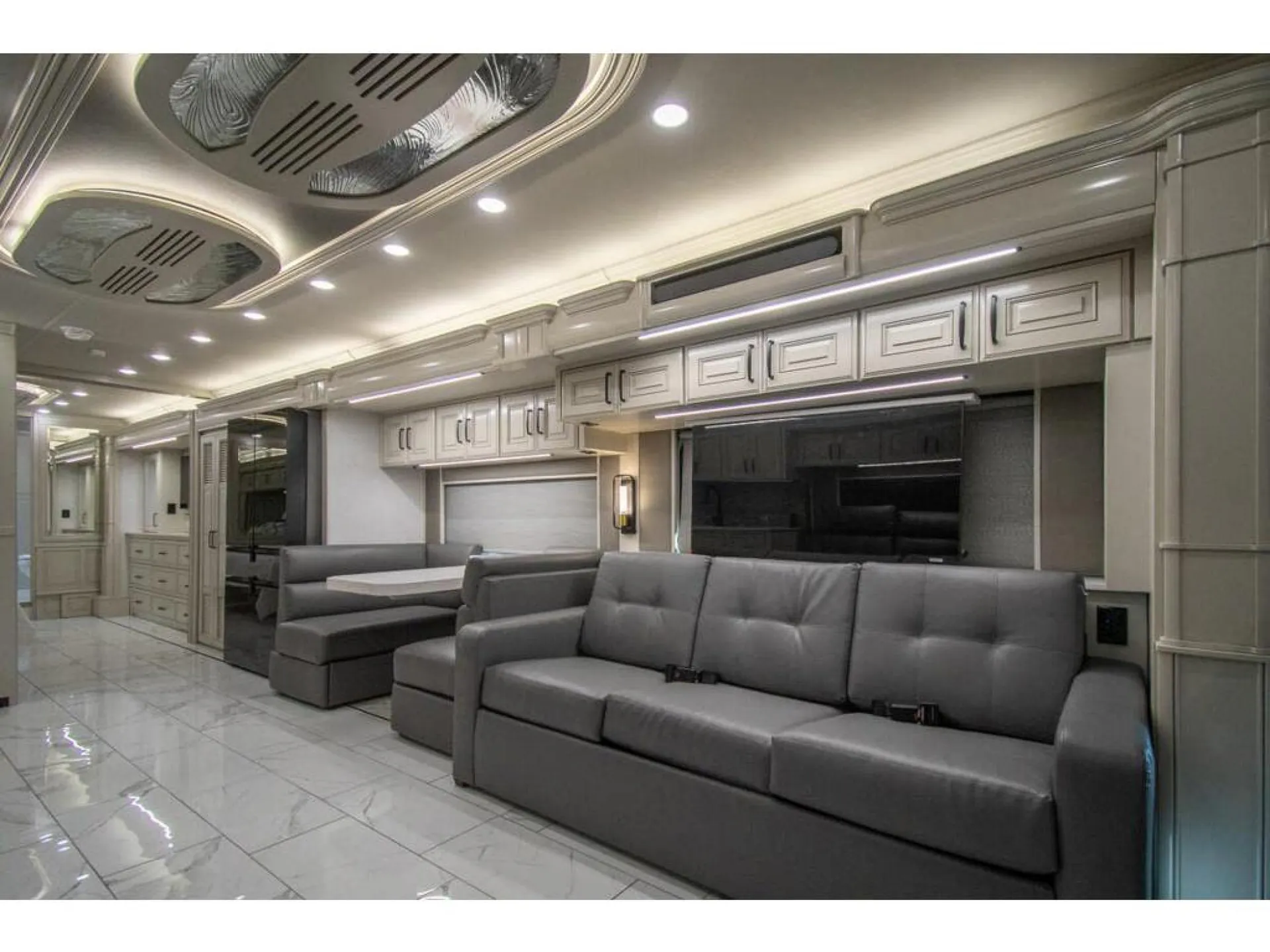 2026 American Coach American Dream 45A — Motorhome living area with a large sofa, opposing lounge seating, entertainment wall, and dinette extending behind it