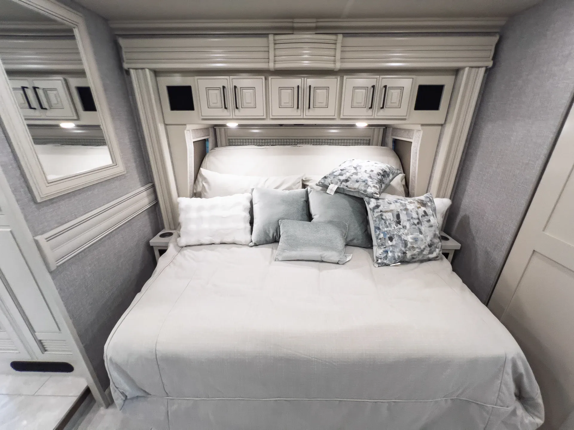 2027 Fleetwood Discovery LXE 40M — Main bedroom with a made bed, pillows, overhead cabinets, bedside surfaces, and a wall mirror