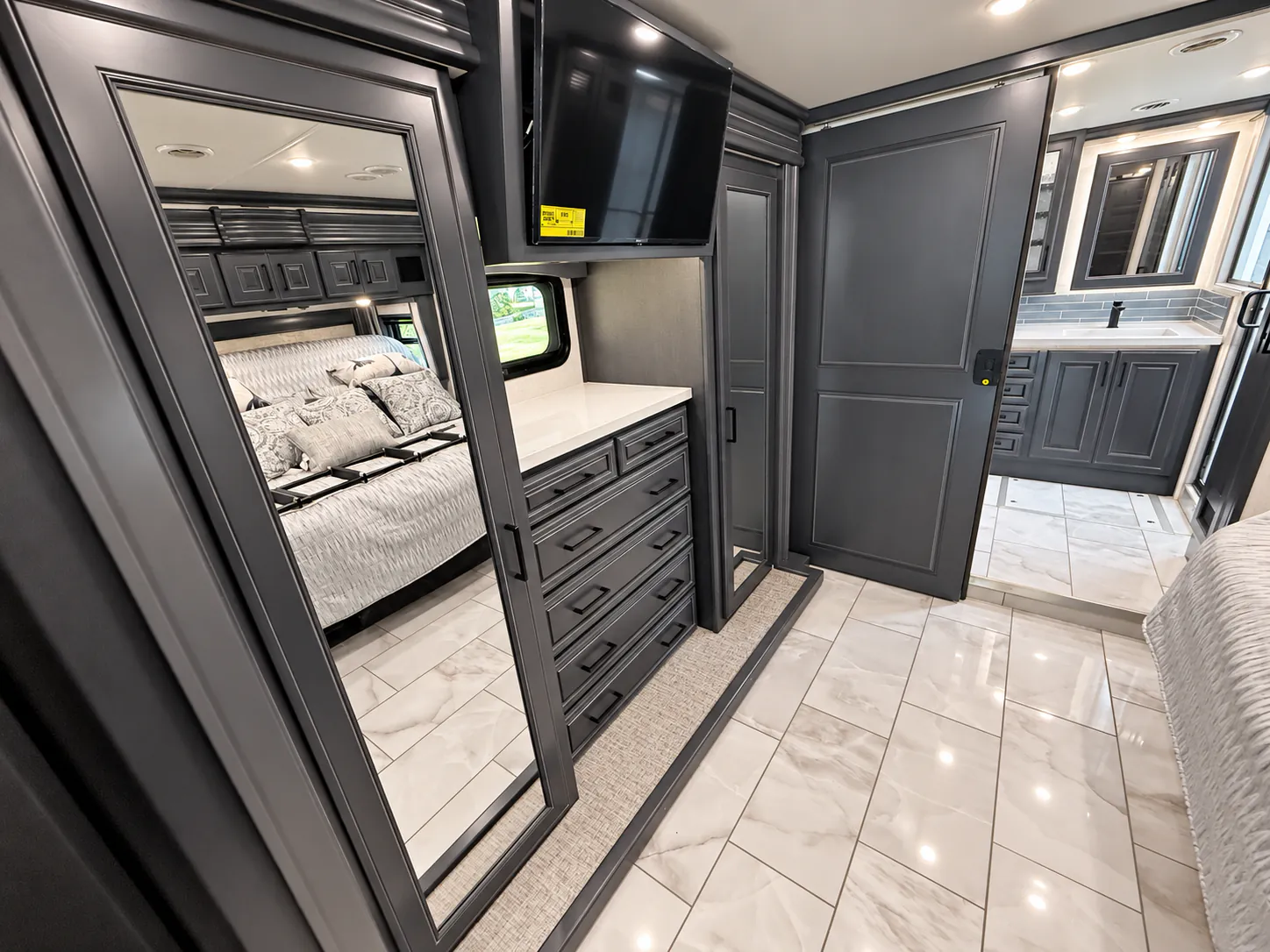 2027 Fleetwood Discovery LXE 40M — Enclosed bedroom with a made bed, mirrored wardrobes, dresser, television, and bathroom vanity visible through the doorway