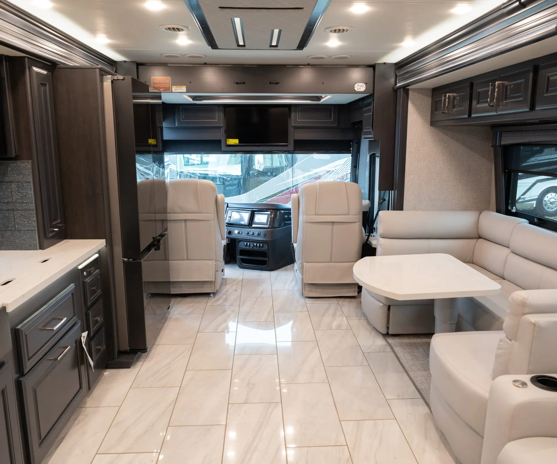 2026 Fleetwood Discovery LXE 40G — Class A front interior overview with cockpit, captain’s chairs, sofa, lounge table, refrigerator, and kitchen cabinetry