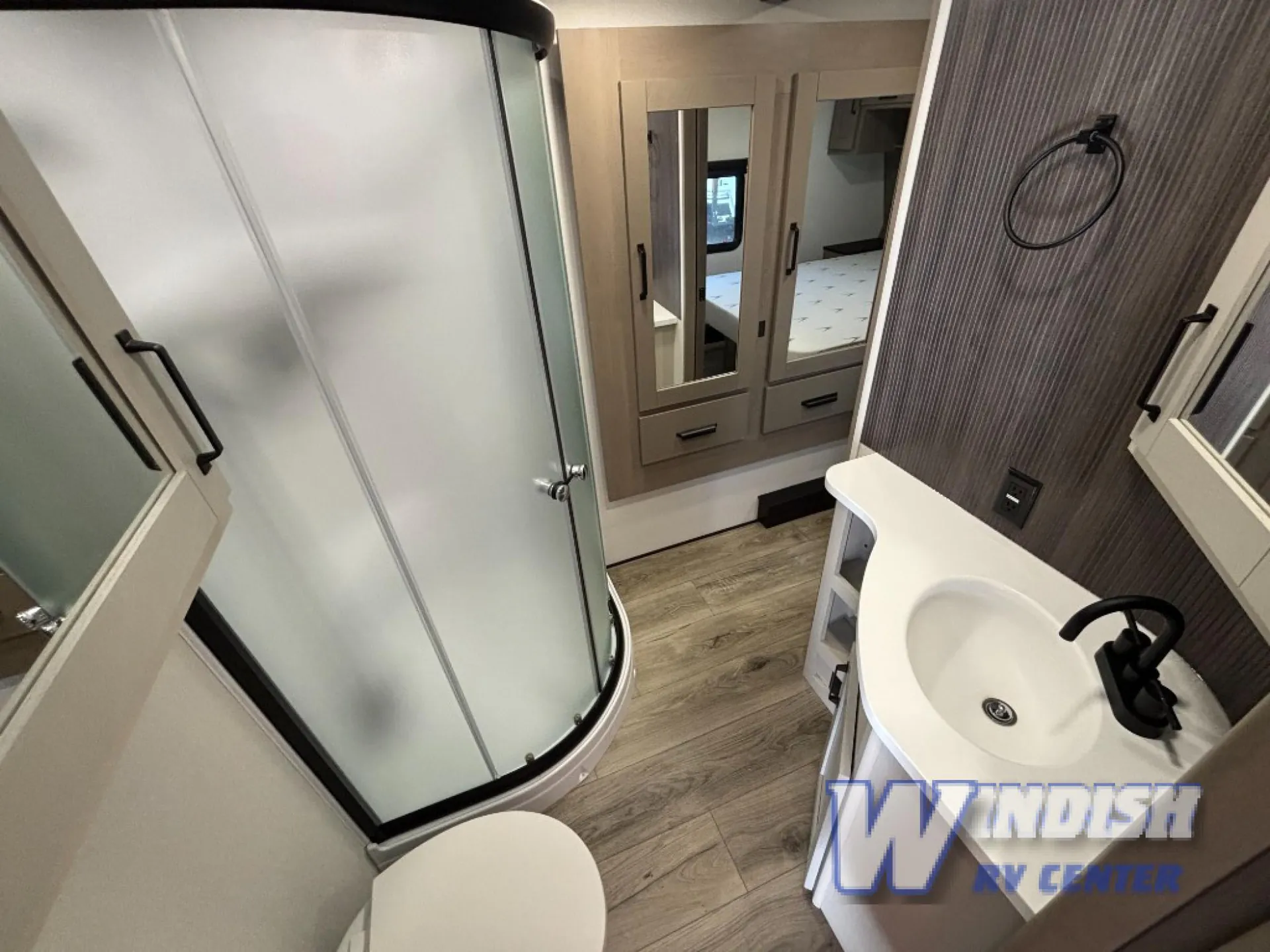 2027 Grand Design Reflection 362TBS — Bathroom showing a corner shower, toilet, curved vanity sink, mirror, and bedroom visible through the doorway