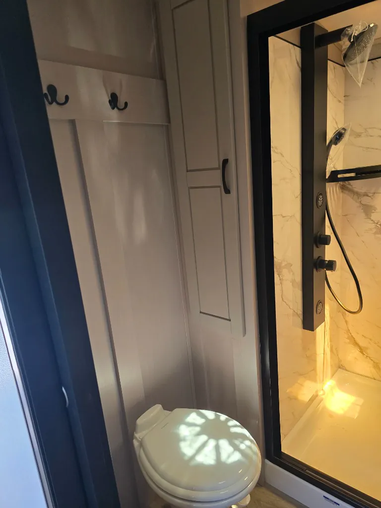 2026 Jayco North Point 377RLBH — Fifth wheel bathroom showing toilet, wall hooks, storage cabinet, and adjacent glass shower enclosure