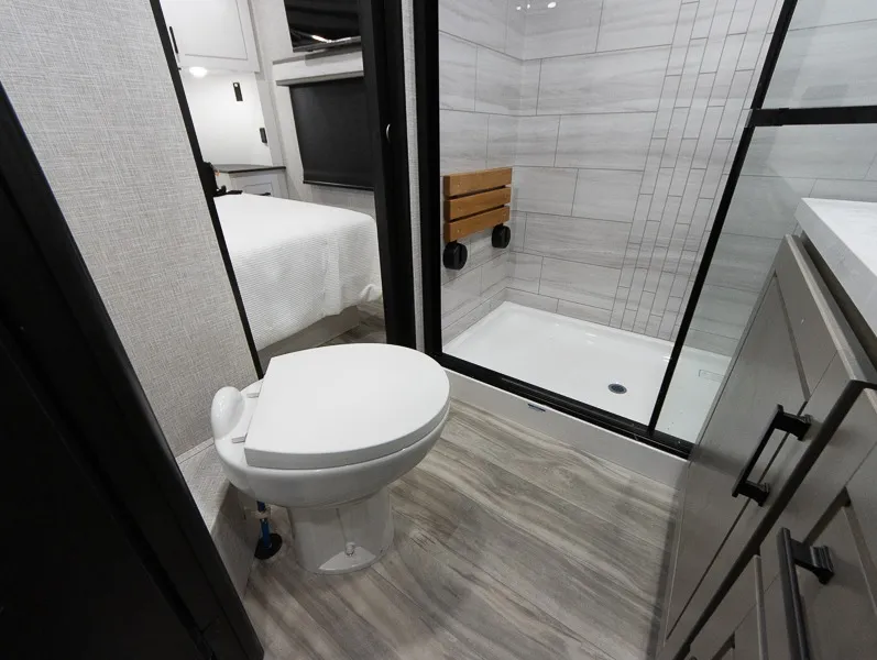 2025 Jayco Seismic Luxury Series 4113 — Bathroom with toilet, tiled shower, and vanity beside a visible bedroom bed through the doorway